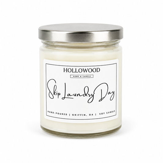 A glass jar candle with a silver metal lid and a white label that reads 'HOLLOWOOD Home & Candle Skip Laundry Day'.