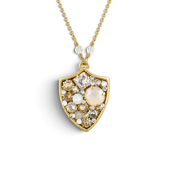 A gold shield pendant necklace featuring a central round pale pink stone surrounded by small clear and iridescent stones, suspended from a delicate gold chain on a white background.