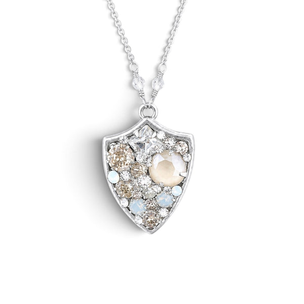 A silver shield-shaped pendant necklace featuring a central cream-colored pearl surrounded by clear, champagne, and light blue accent crystals, suspended from a delicate silver chain on a white background.